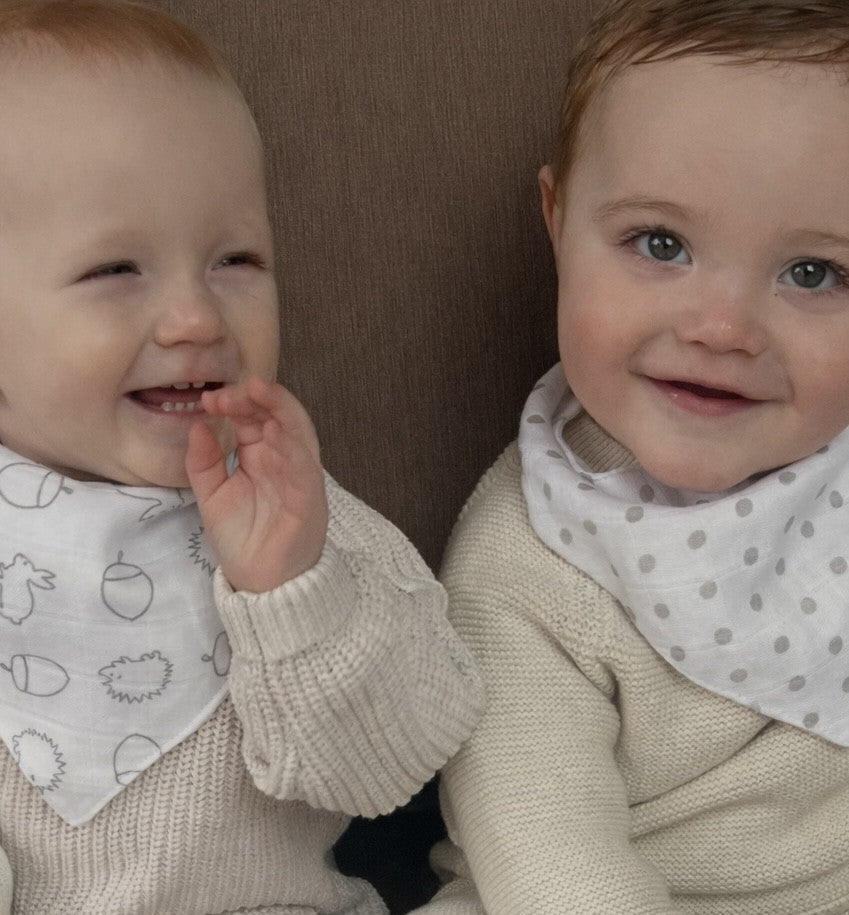 Two babies wearing beige sweaters and bibs sitting next to each other.
